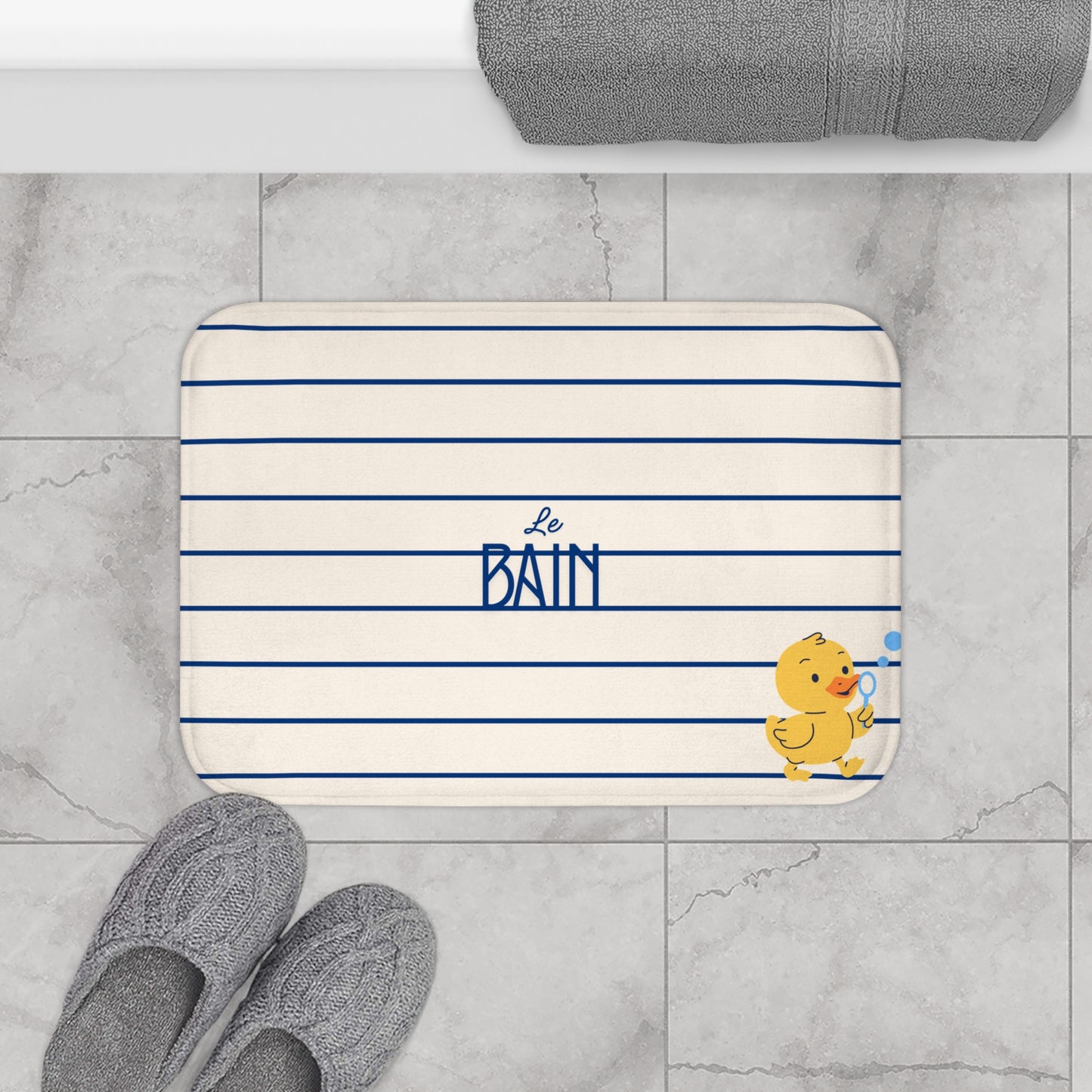 A rectangular bathroom mat with rounded corners, featuring navy blue horizontal stripes on an off-white background, with the text 'Le BAIN' in navy blue and a small yellow duck character holding a blue towel. The mat is styled in a bathroom setting on gray marble tiles, with gray slippers in the foreground and a folded gray towel at the top.