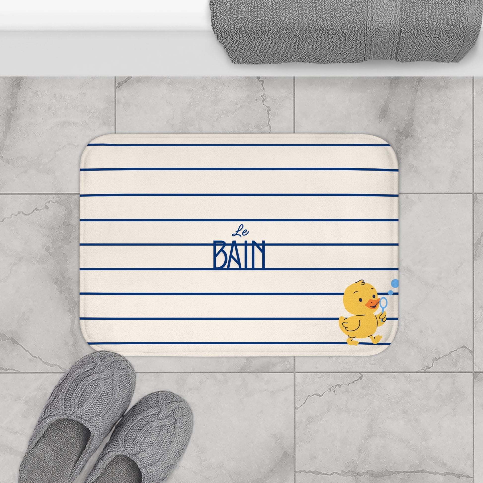 A rectangular bathroom mat with rounded corners, featuring navy blue horizontal stripes on an off-white background, with the text 'Le BAIN' in navy blue and a small yellow duck character holding a blue towel. The mat is styled in a bathroom setting on gray marble tiles, with gray slippers in the foreground and a folded gray towel at the top.