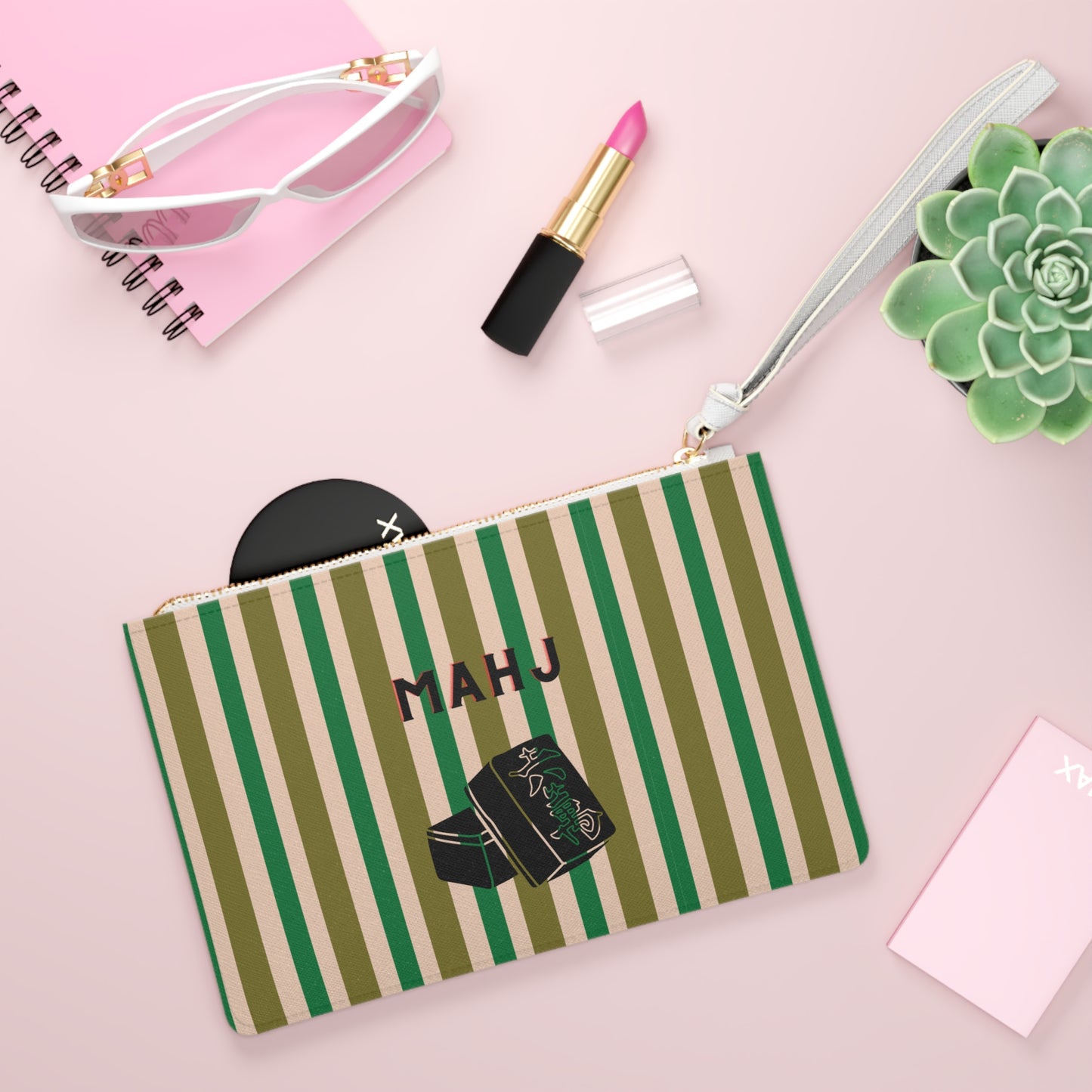 A personalized cosmetic pouch with green and beige vertical stripes, featuring the name 'MAHJ' and a lipstick illustration. The pouch is styled in a flat-lay arrangement with sunglasses, lipstick, a succulent, and a notebook on a pink background.