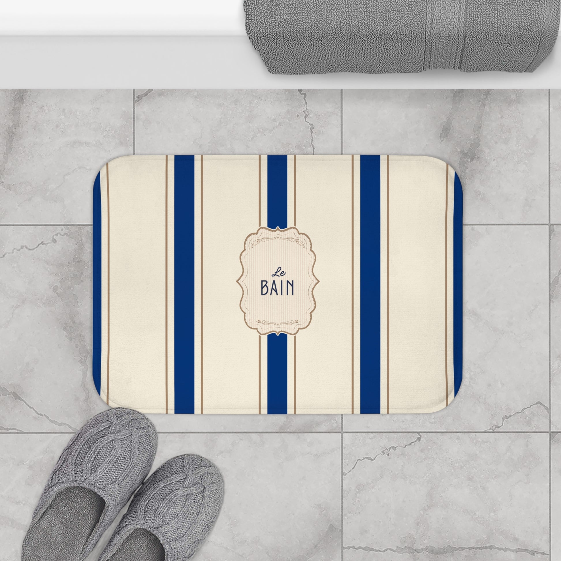 Bath mat with blue stripes and 'Le Bain' text on a marble floor with slippers and towel.