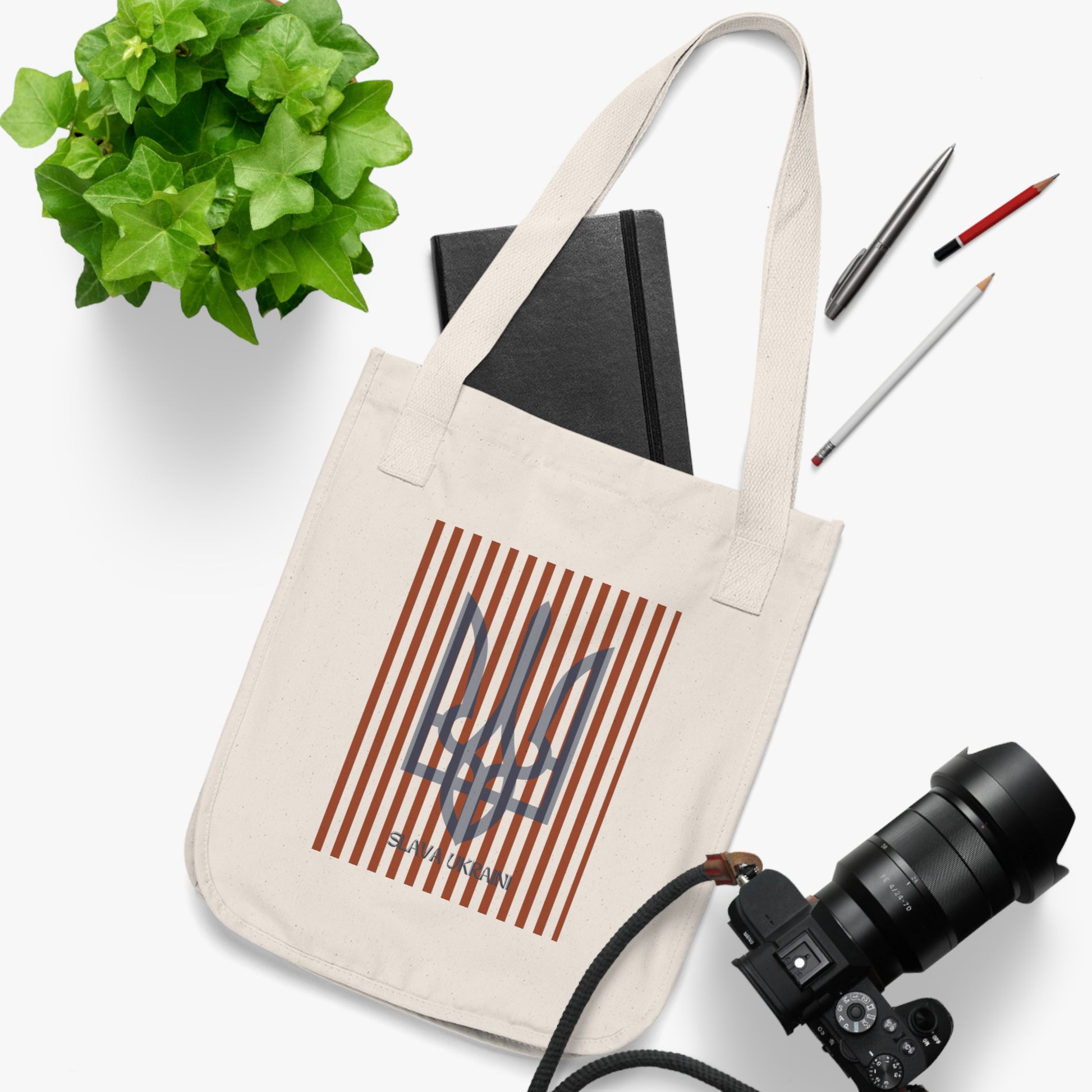 Beige tote bag with a design on a white surface with a plant, notebook, and camera.