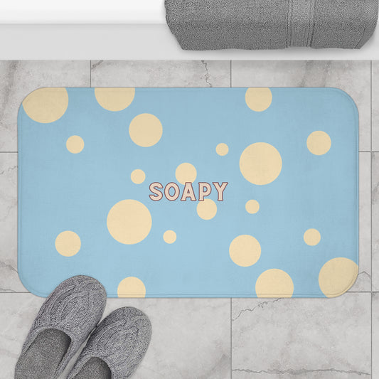 A rectangular blue bath mat with a pattern of large and small yellow circles, personalized with the word 'SOAPY' in the center. The mat is shown on a light gray marble tile floor, with gray slippers at the bottom left and a folded gray towel at the top.