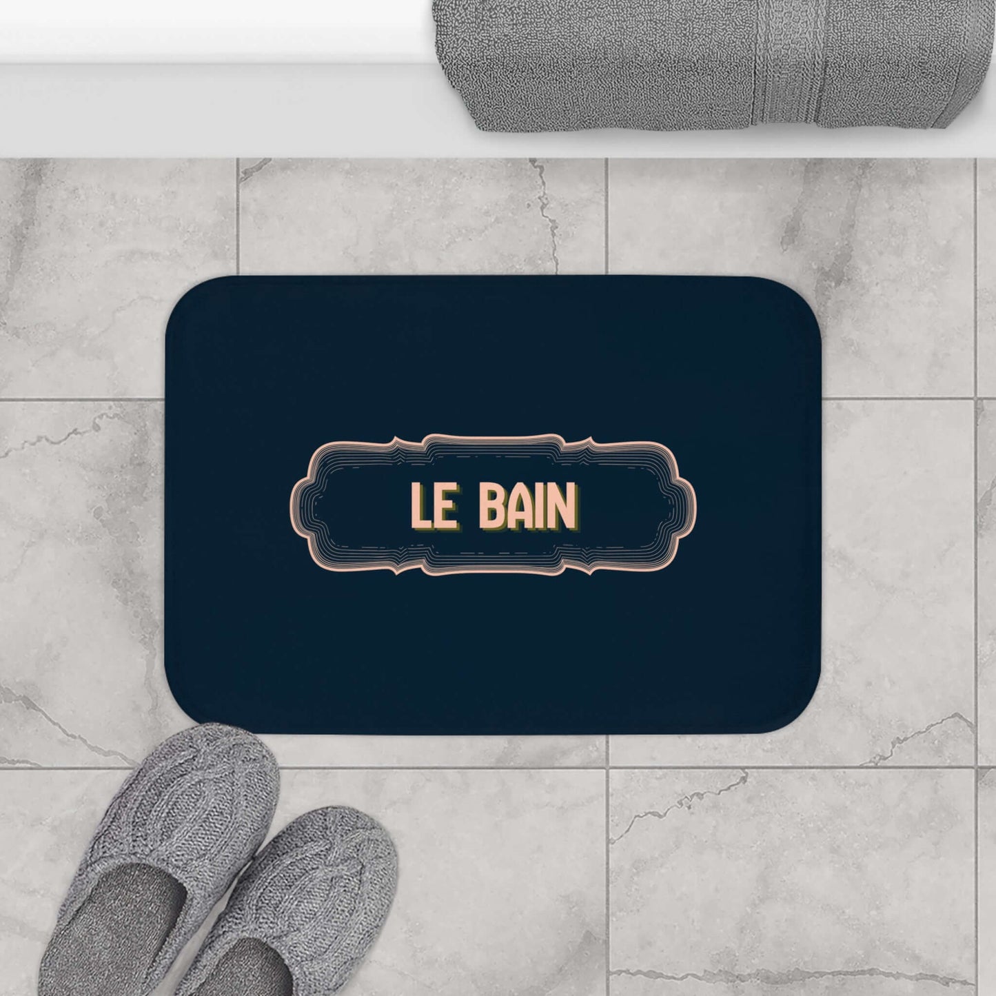 Navy bath mat with 'LE BAIN' text on a marble floor with slippers and a towel.