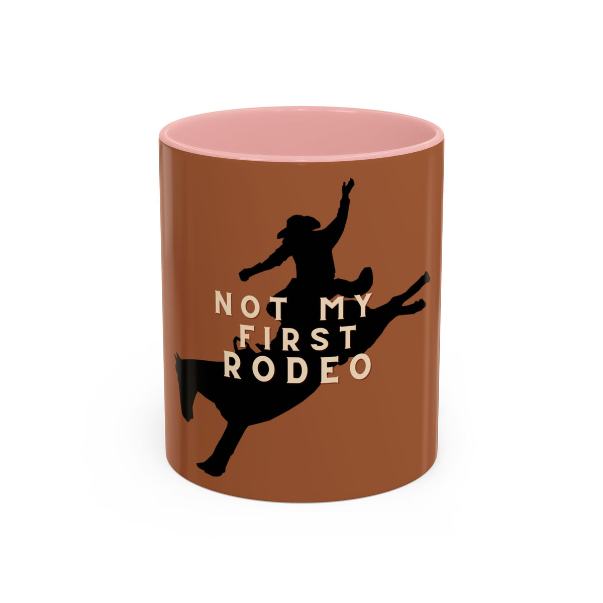 A brown ceramic mug with a pink interior, featuring a black silhouette of a cowboy riding a bucking bull and the text 'NOT MY FIRST RODEO' in beige.