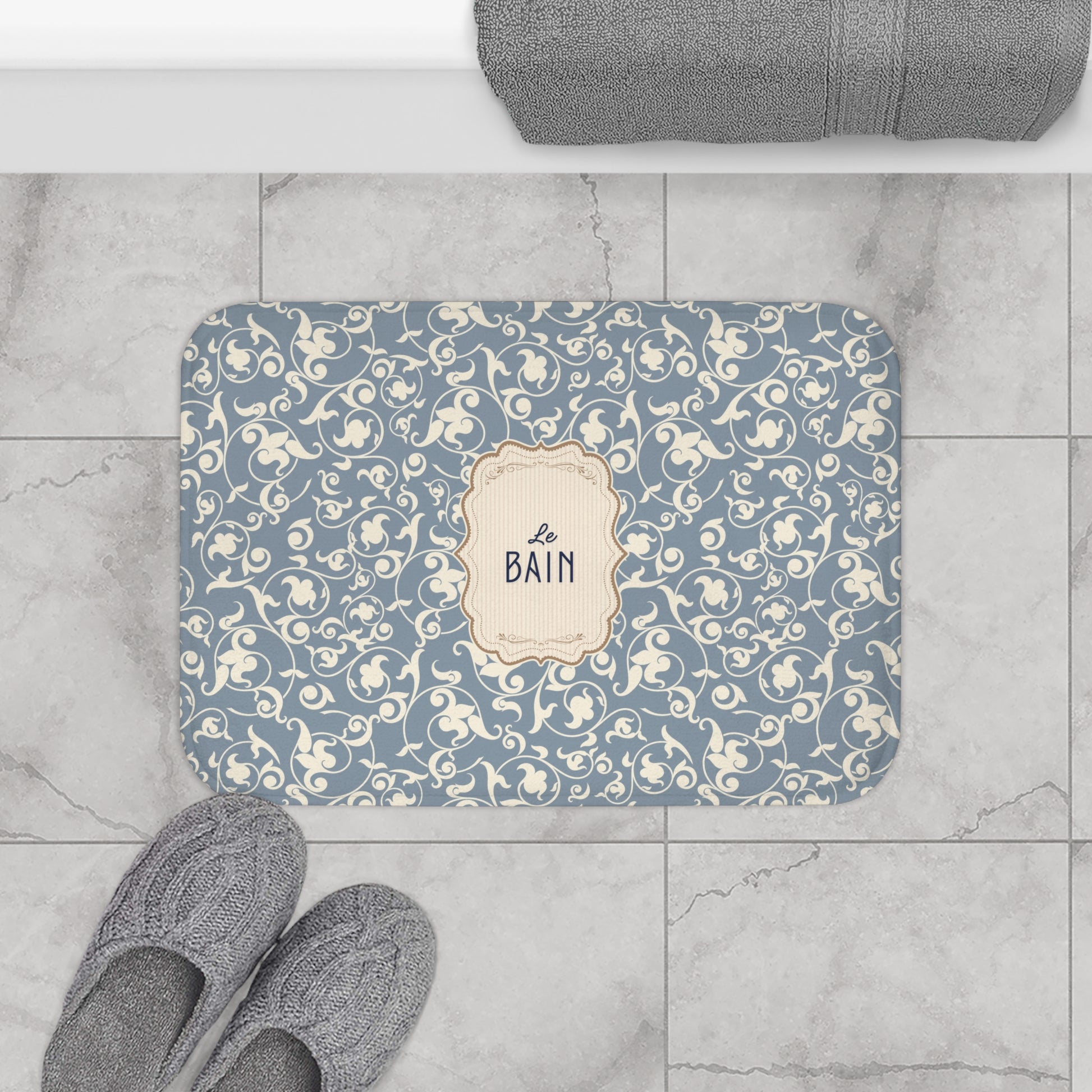 Decorative bath mat with floral pattern and 'Le Bain' text on a tiled floor.