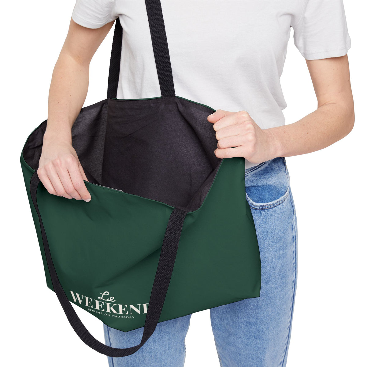 A person wearing a white t-shirt and blue jeans holds a dark green tote bag with black handles. The bag features the text 'The WEEKENI' in white on the front, with a black interior lining.