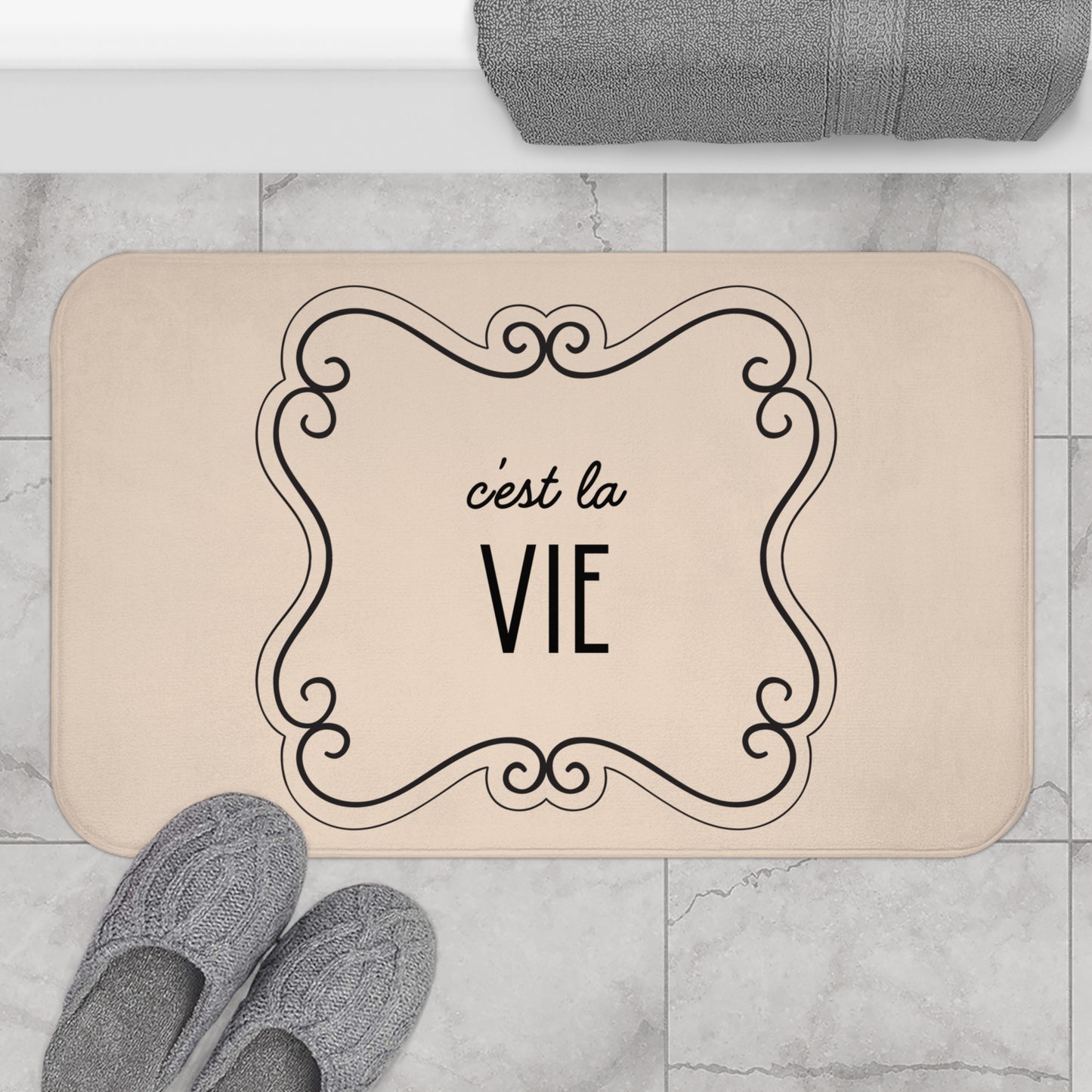 Beige doormat with 'c'est la Vie' text on a marble floor with slippers and a towel.