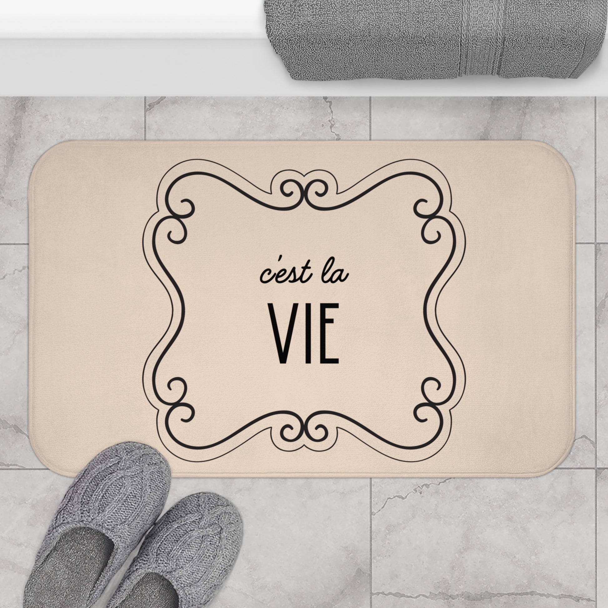 Beige doormat with 'c'est la Vie' text on a marble floor with slippers and a towel.