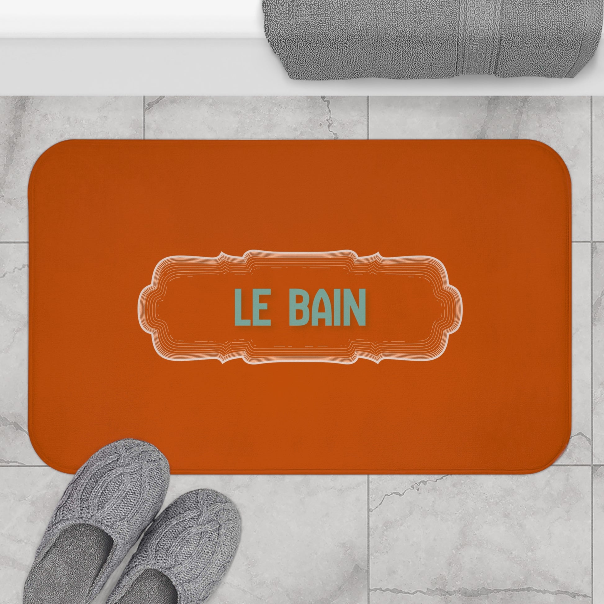 Orange bath mat with 'LE BAIN' text on a marble floor with slippers and a towel.
