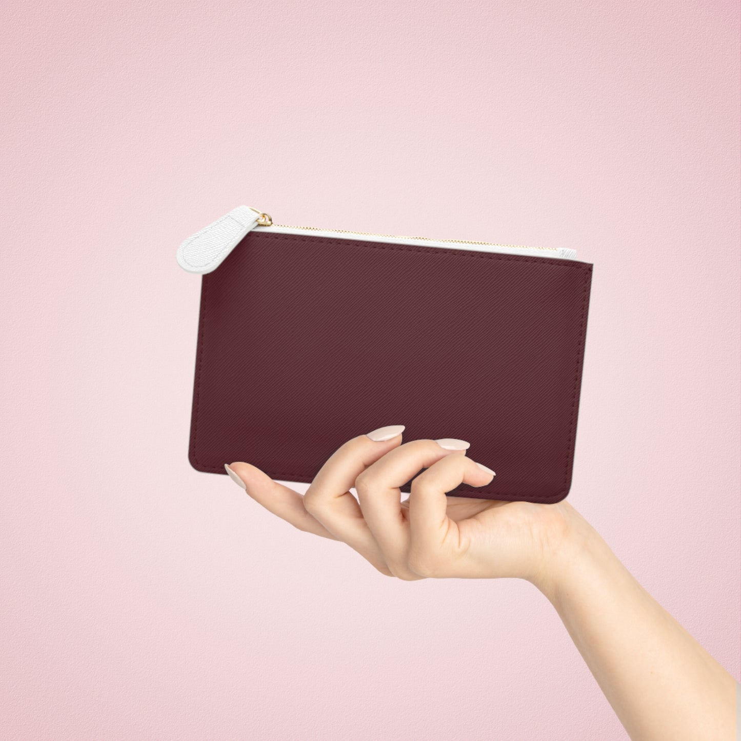 Hand holding a burgundy pouch with a white zipper on a pink background