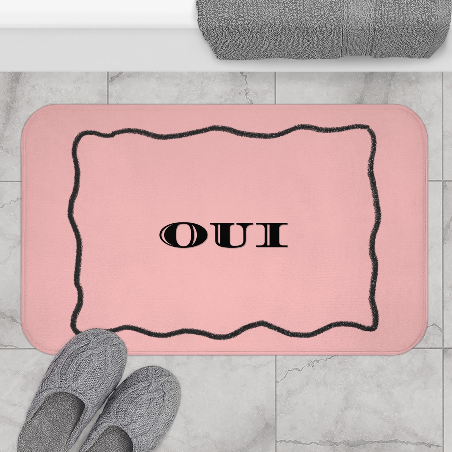 Pink bath mat with 'OUI' text on a marble floor with slippers and a towel.