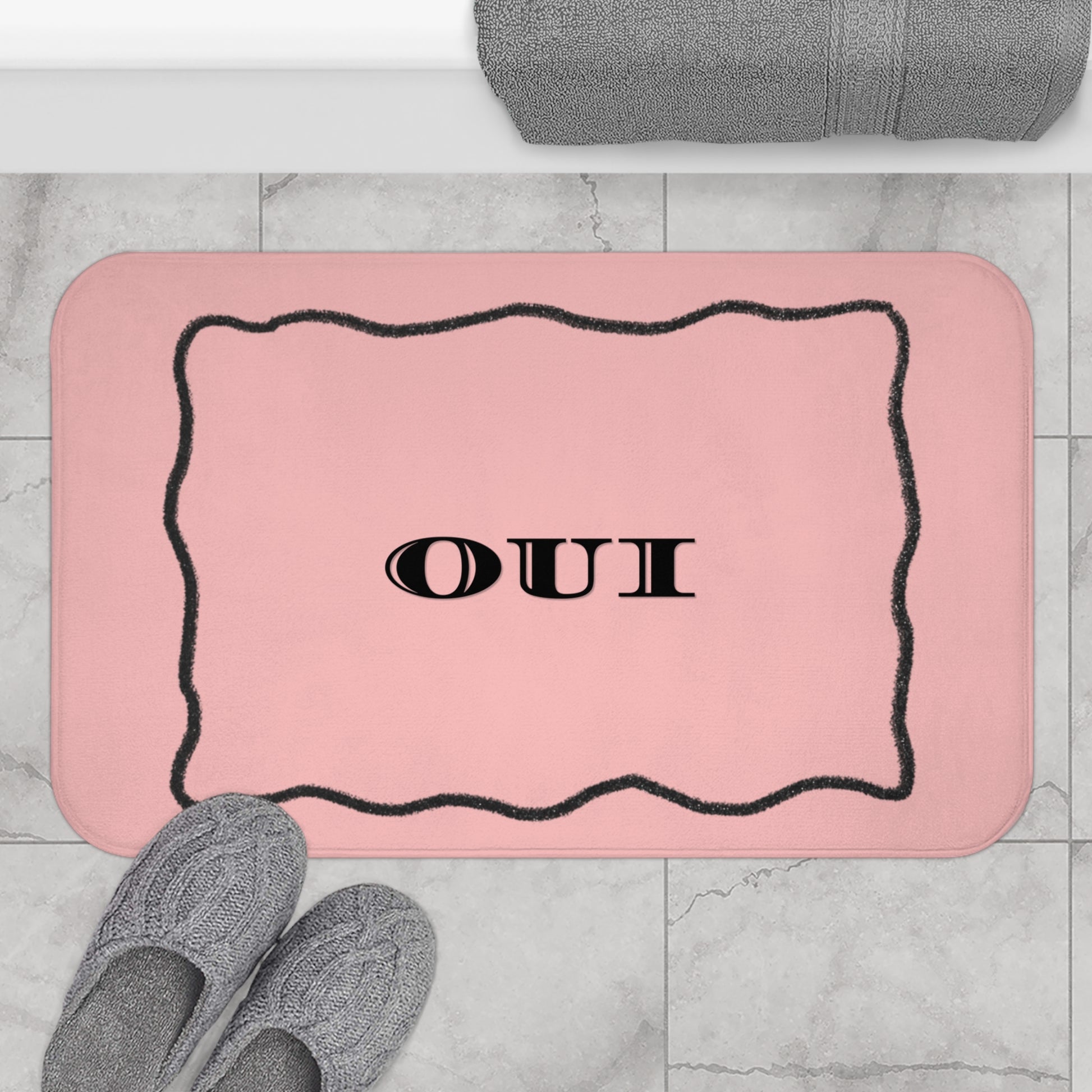 Pink bath mat with 'OUI' text on a marble floor with slippers and a towel.