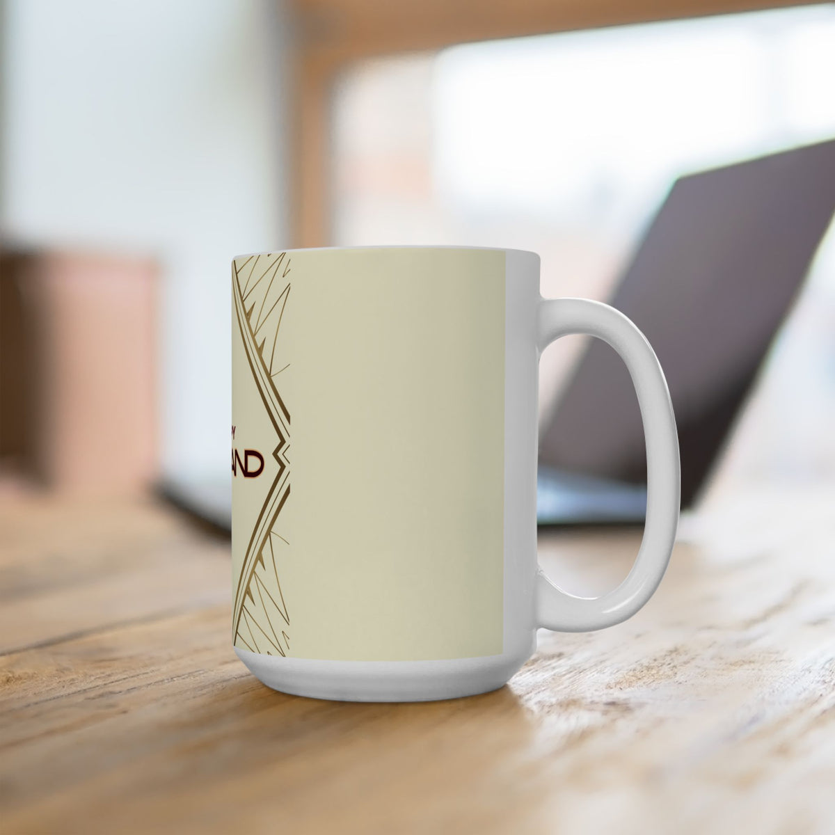 A white ceramic coffee mug with a gold and black typographic design sitting on a wooden table in a blurred indoor setting with a laptop in the background.