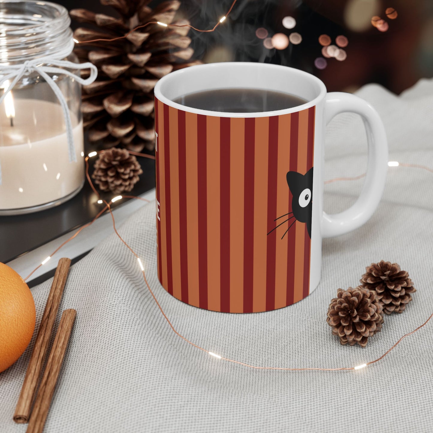 A white ceramic mug with a red and orange striped pattern and a black cat face graphic, filled with a hot beverage and set on a table with pinecones, fairy lights, a candle, and cinnamon sticks.