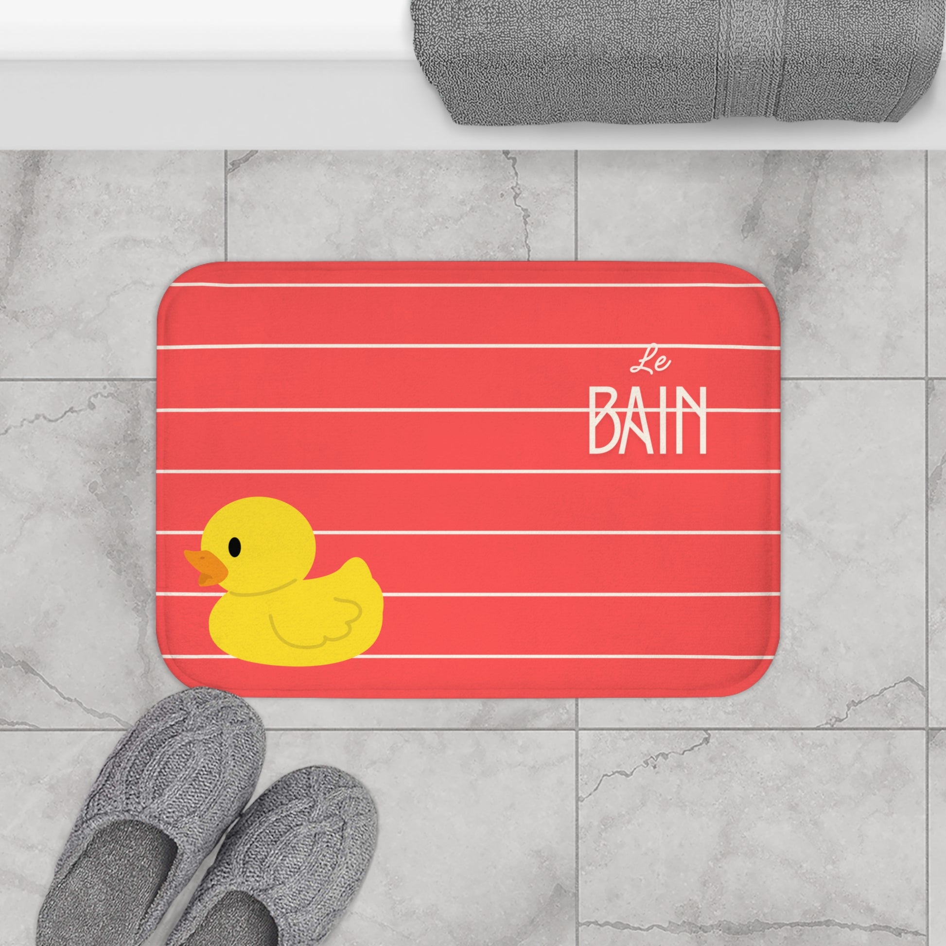 Bath mat with a rubber duck and 'Le Bain' text on a red background, placed on a tiled floor.