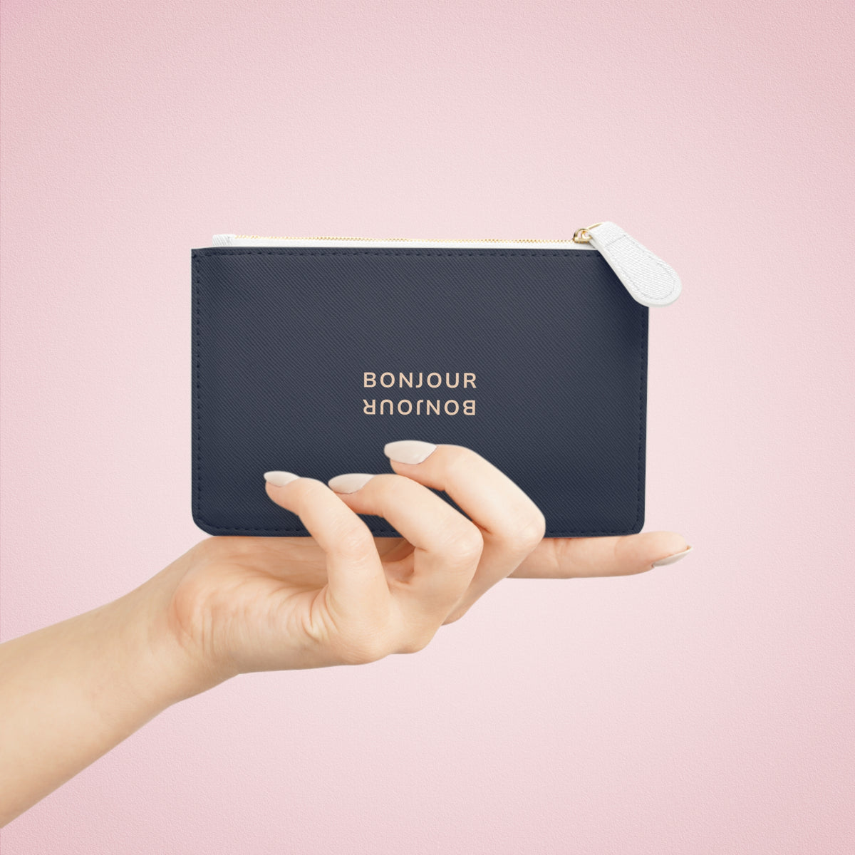 A navy blue leather zippered pouch with gold foil 'BONJOUR' lettering, held by a hand against a solid pink background.