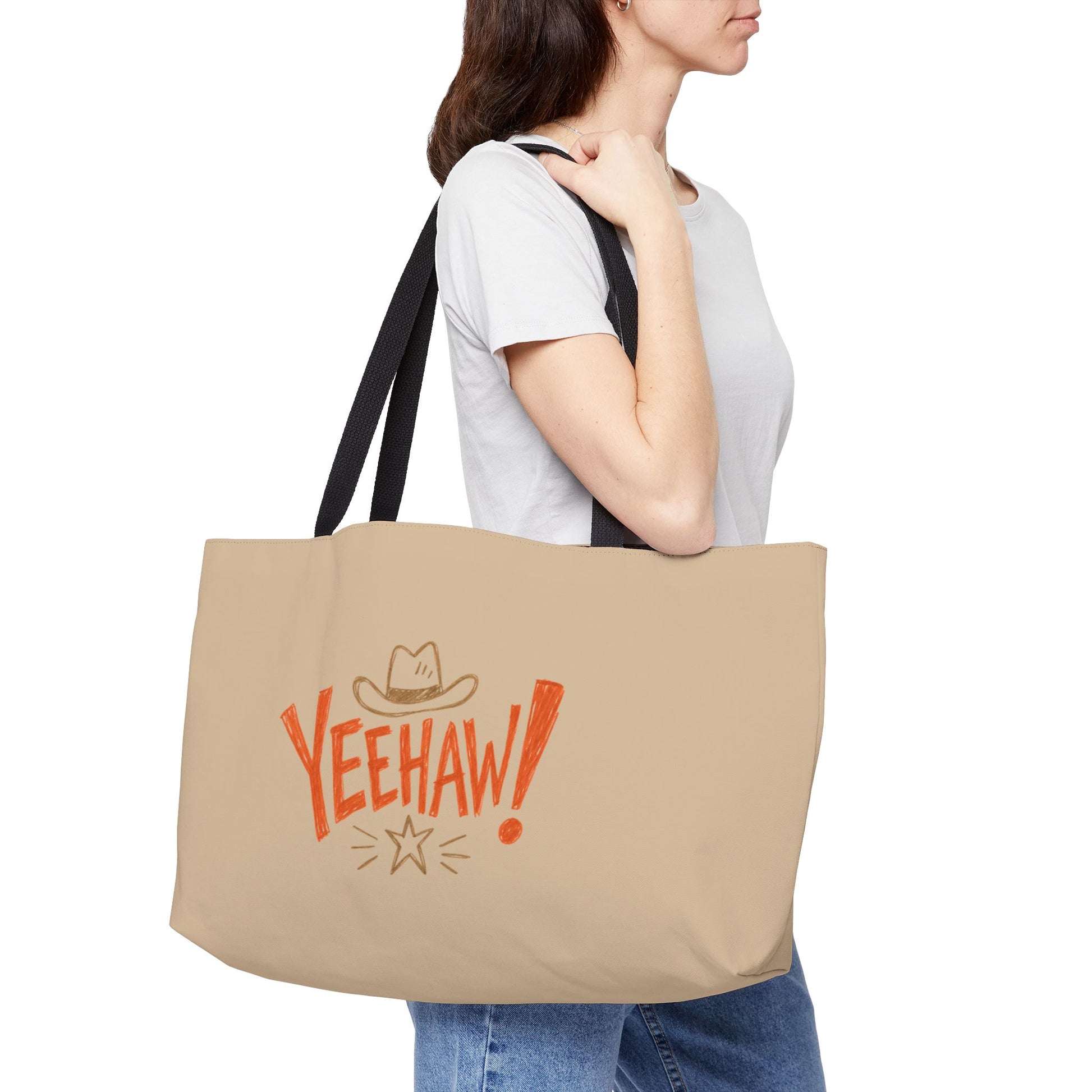 A woman wearing a white t-shirt and blue jeans carries a beige canvas tote bag over her shoulder. The bag features black straps and a western-themed graphic with the word 'YEEHAW!' in orange, a cowboy hat, and a star.