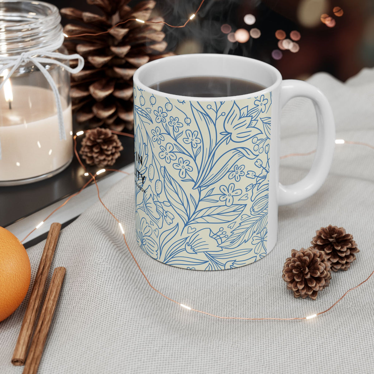 White mug with hot chocolate, surrounded by decorative elements like a candle, cinnamon sticks, and pine cones on a table.