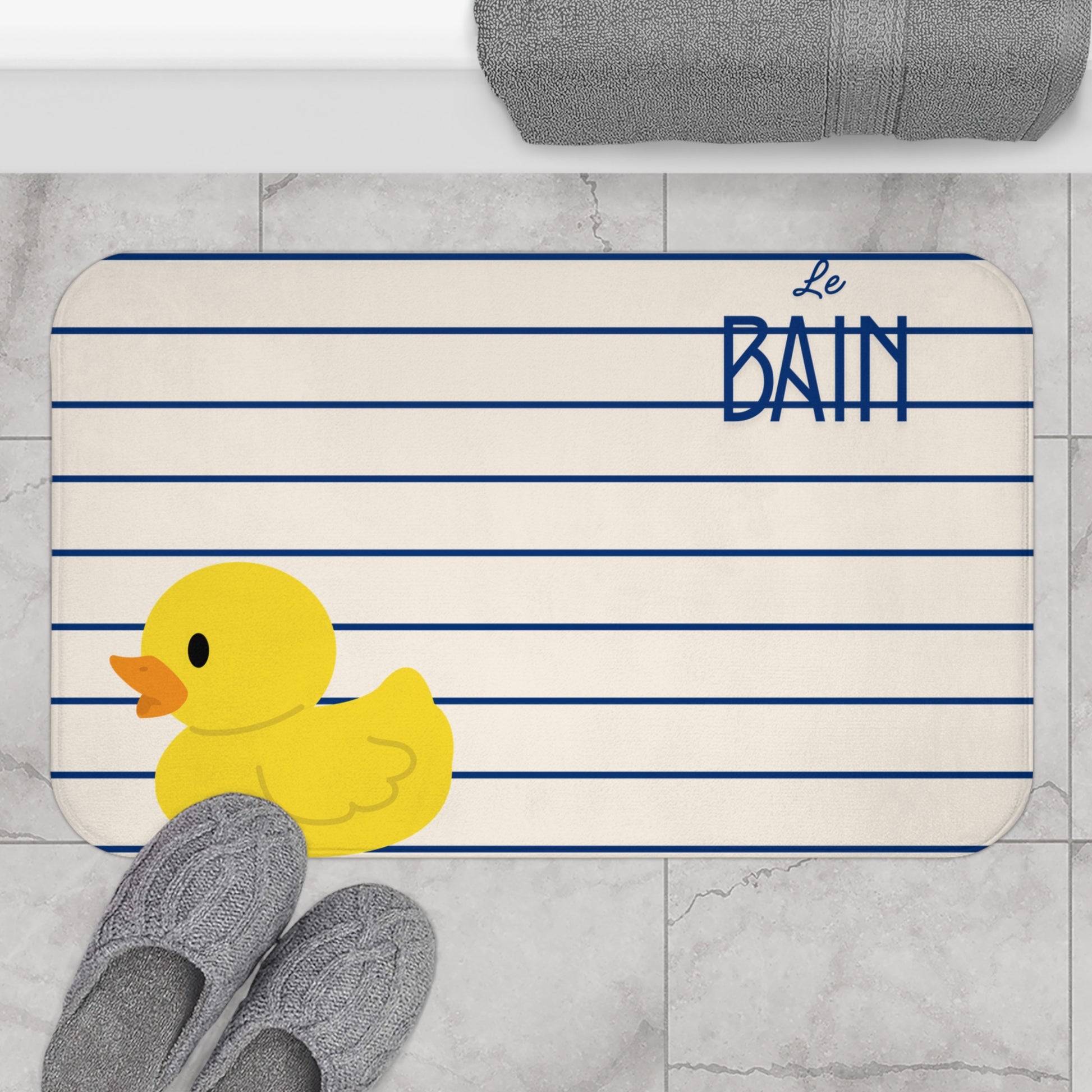 Bath mat with a yellow rubber duck and 'Le Bain' text on a striped background, placed on a tiled floor.