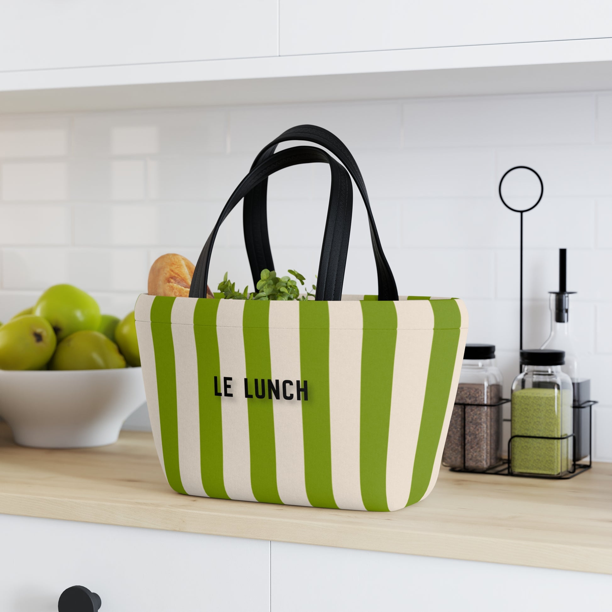 Green and white striped bag labeled 'LE LUNCH' on a kitchen counter with fruits and kitchen items in the background.