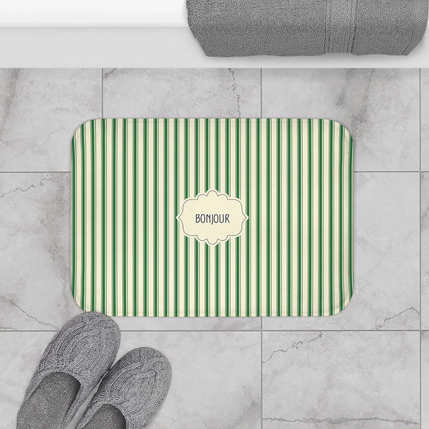 A rectangular bath mat with green and white vertical stripes and a cream label in the center that reads 'BONJOUR', shown on a light marble tile floor with gray slippers and a folded gray towel nearby.