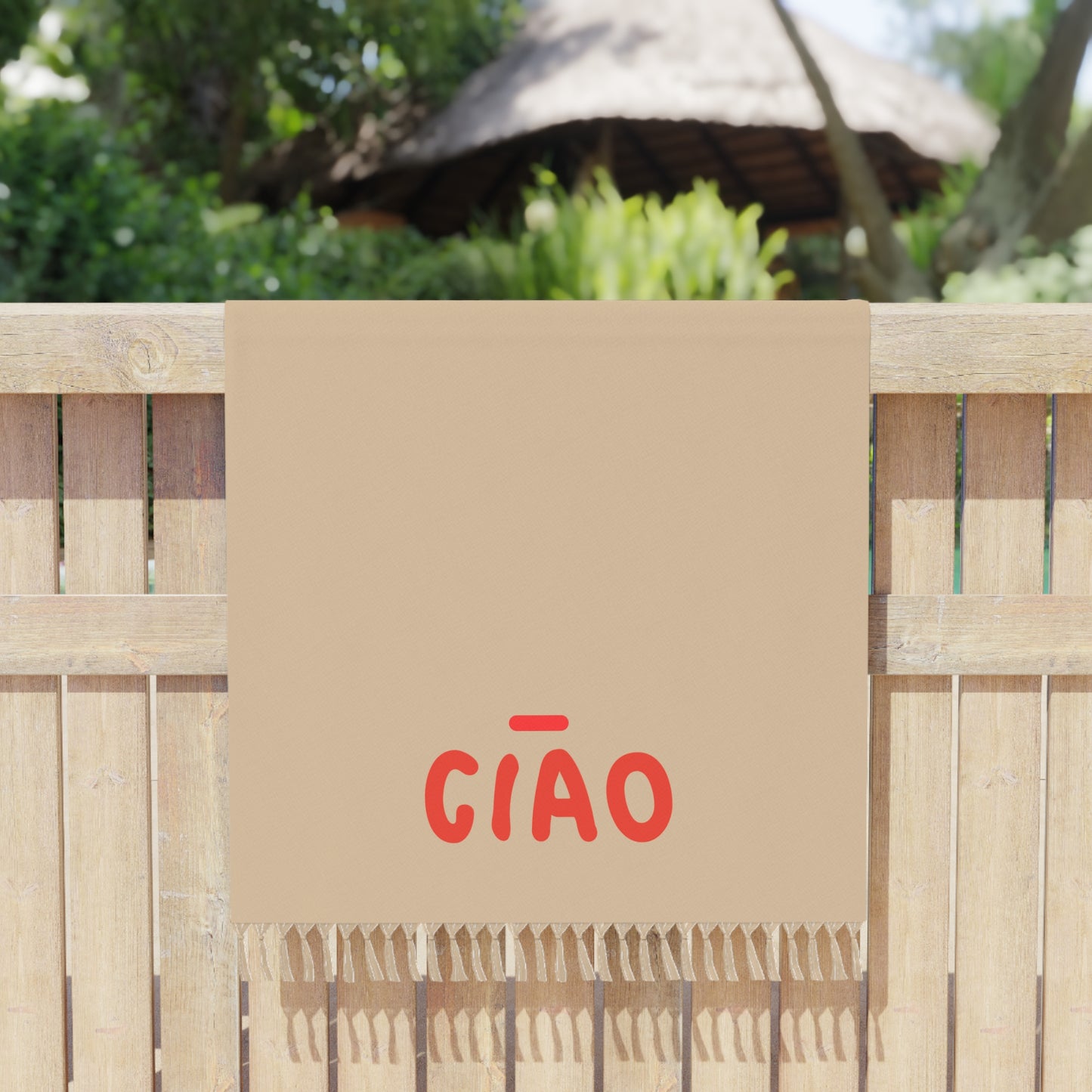 Beige towel with red 'GIÓO' text draped over a wooden fence with a thatched roof structure in the background.