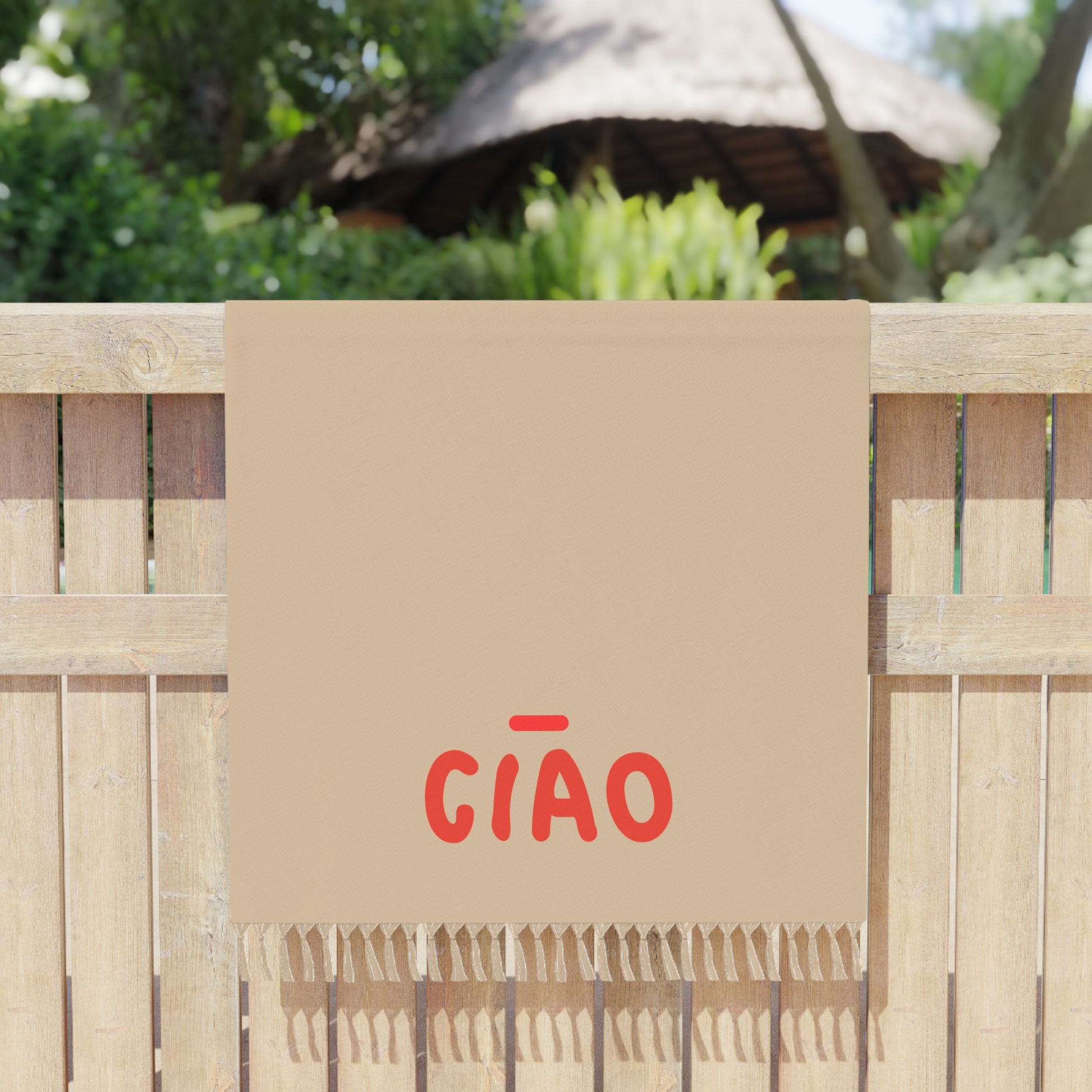 Beige towel with red 'GIÓO' text draped over a wooden fence with a thatched roof structure in the background.