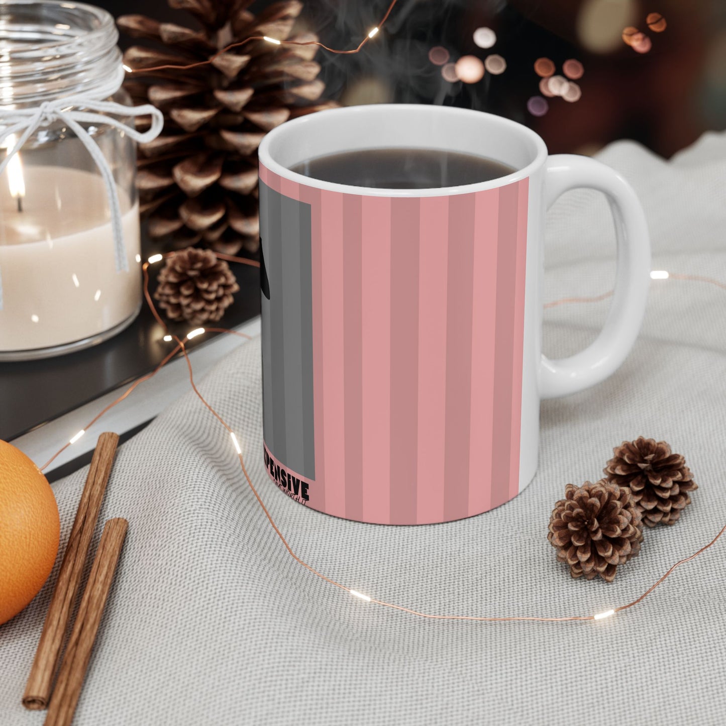 A white ceramic mug filled with a dark beverage, featuring vertical pink and gray stripes on the side, styled on a light fabric surface with pinecones, a candle jar, cinnamon sticks, and fairy lights.