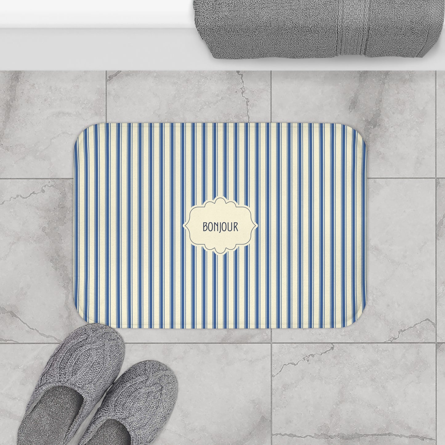 A rectangular bath mat with blue and cream vertical stripes and a central cream label reading 'BONJOUR', shown on a light marble tile floor with gray towels and slippers nearby.