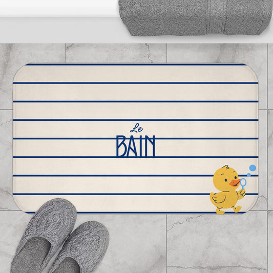 A personalized bath mat with navy blue horizontal stripes, the text 'Le BAIN' in navy blue, and a yellow cartoon duck holding a blue bubble wand. The mat is styled on a light gray marble tile floor with gray knit slippers and a folded gray towel visible at the top.