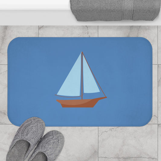 A rectangular blue bath mat with rounded corners featuring a stylized sailboat illustration, shown on a light marble floor with gray slippers and a folded gray towel nearby.