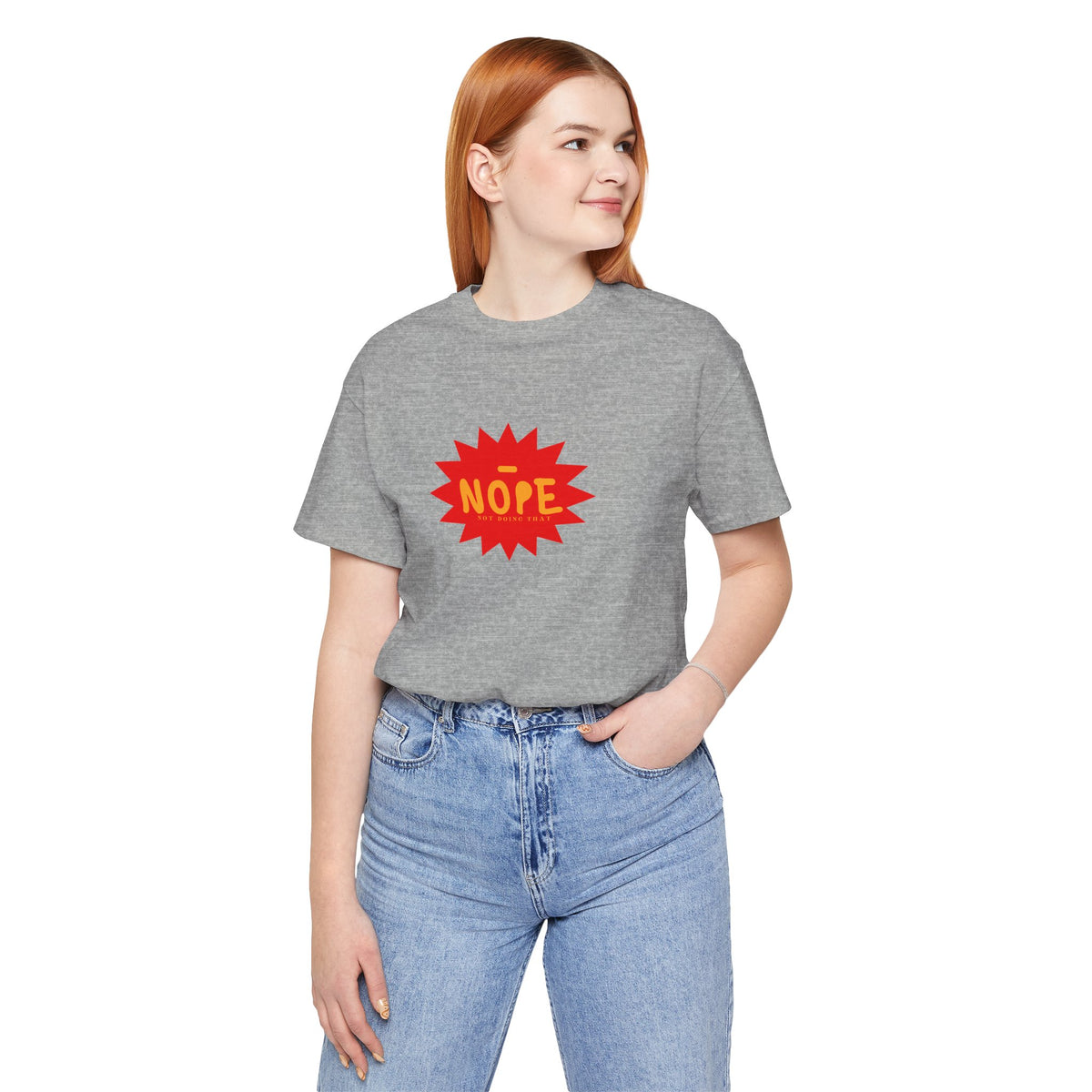 A woman with red hair wearing a heather gray t-shirt with a red starburst graphic that says 'NOPE' in yellow, paired with light blue jeans, against a plain white background.