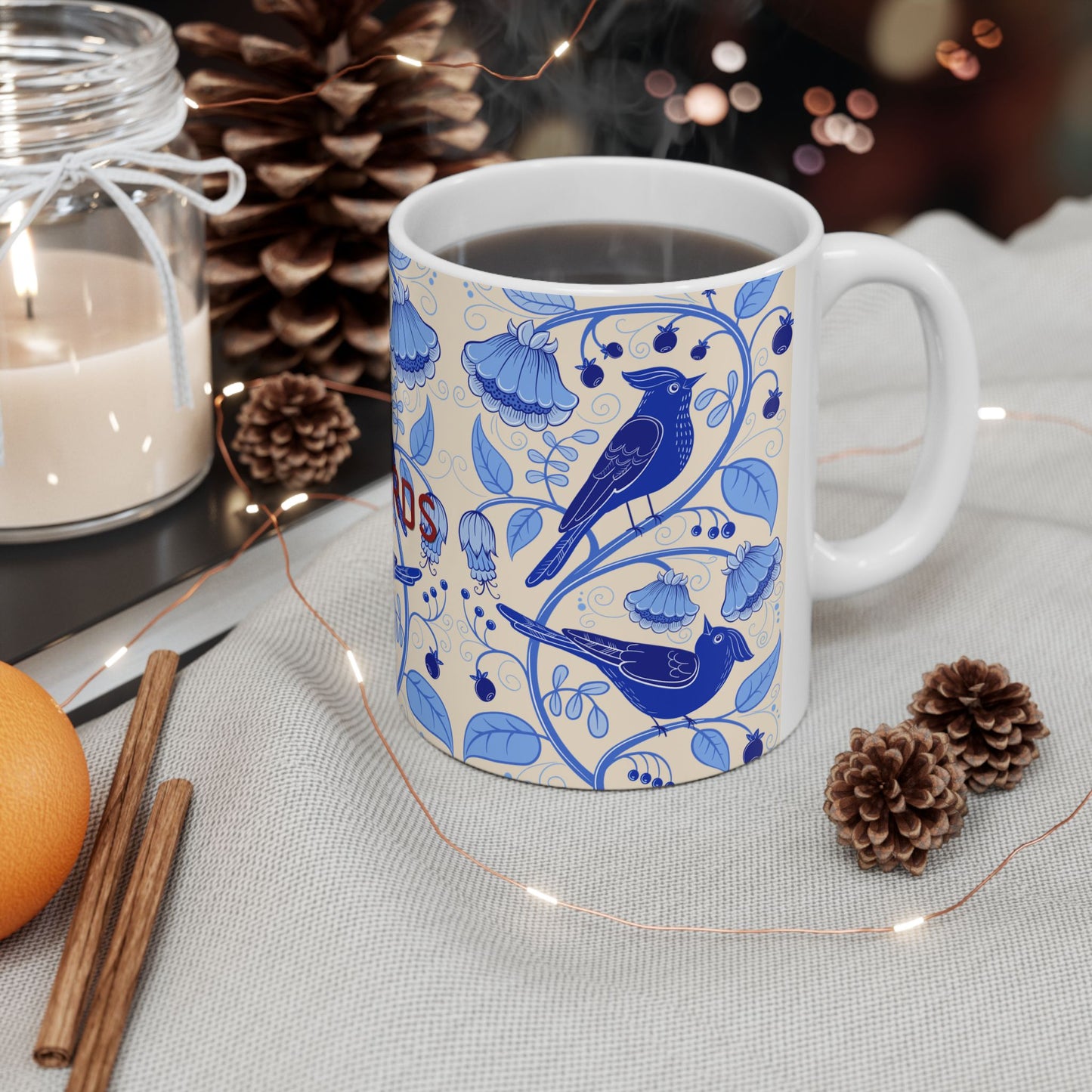 A white ceramic coffee mug with a blue floral and bird pattern, filled with black coffee and set on a light fabric surface with pinecones, a candle, cinnamon sticks, and fairy lights.