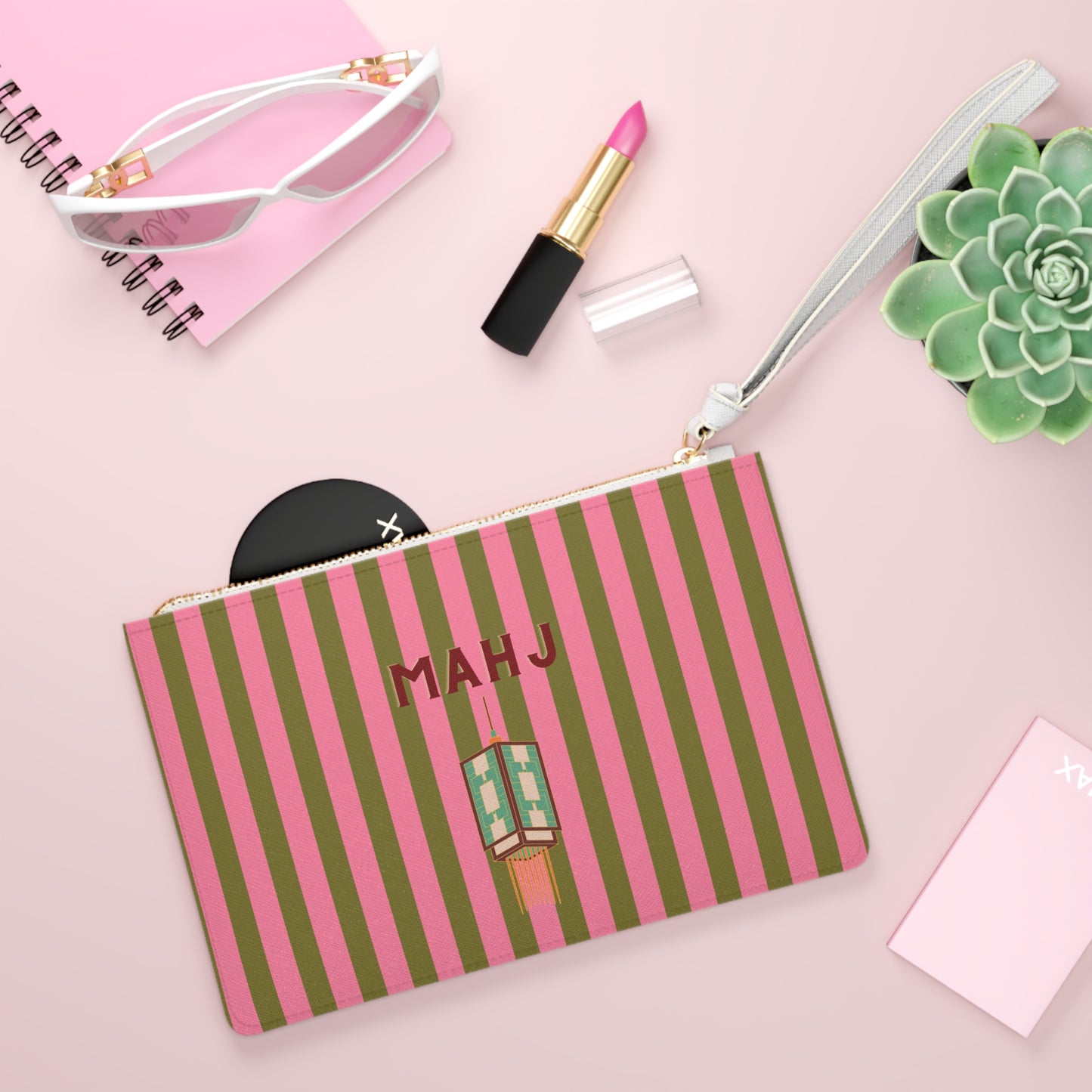 A pink and olive green striped cosmetic pouch with a wrist strap, featuring the name 'MAHJ' and a lipstick illustration on the front. The pouch is styled in a flat lay with pink sunglasses, a pink notebook, a pink lipstick, a succulent, and a black compact on a light pink background.