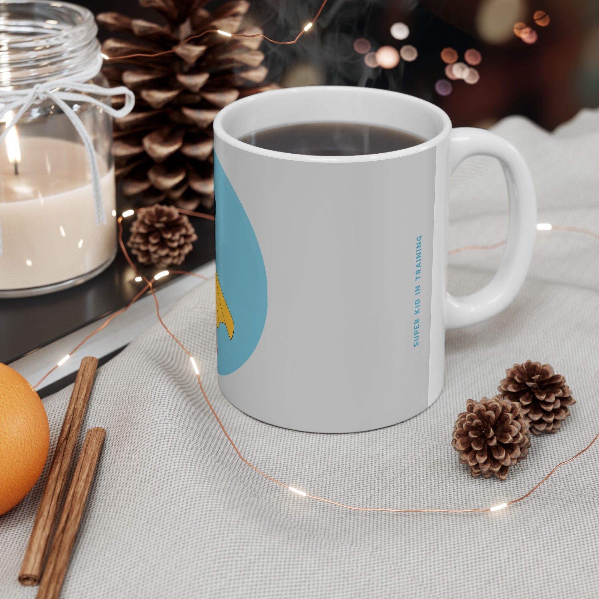 A white ceramic coffee mug with a light blue and yellow graphic design, filled with a hot beverage and set on a textured tablecloth surrounded by cozy holiday props like pinecones, a candle, cinnamon sticks, and fairy lights.