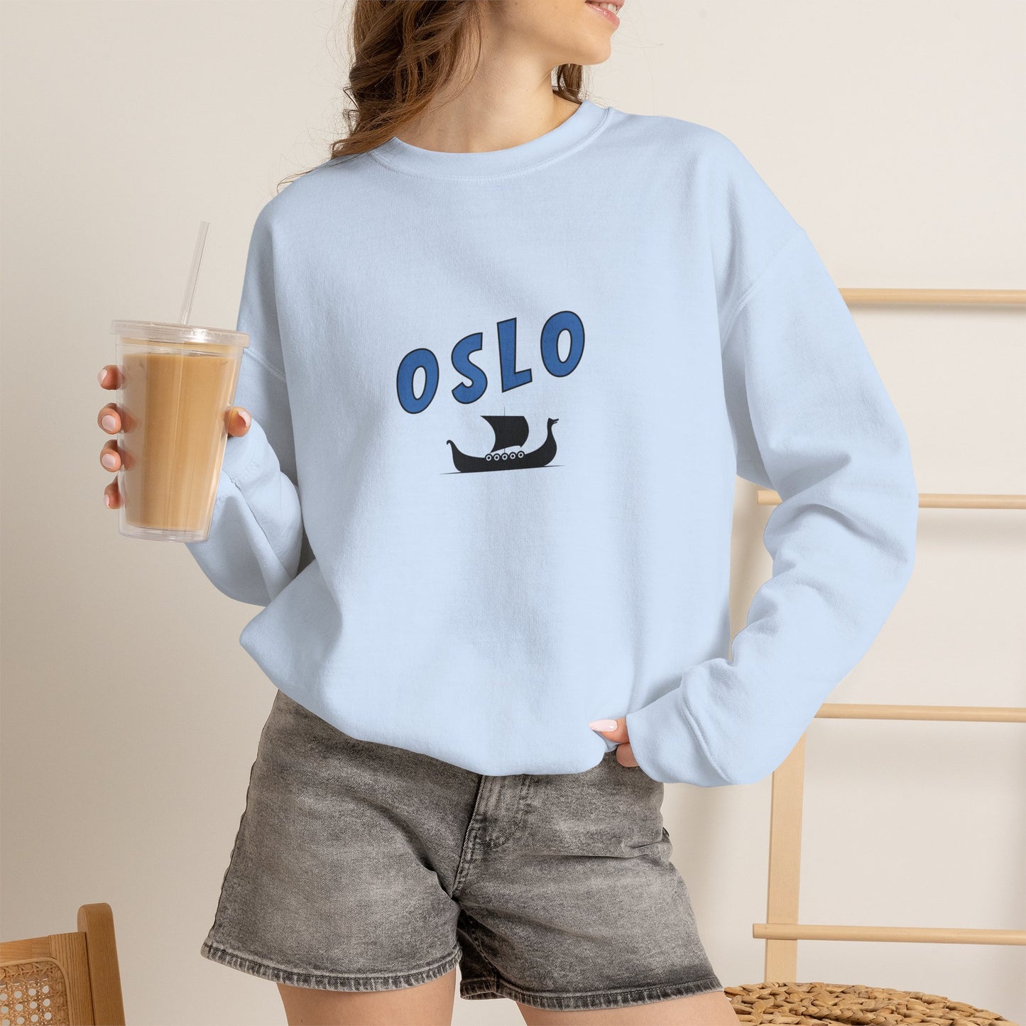 Person wearing a light blue sweatshirt with 'OSLO' and a ship graphic, holding a drink.