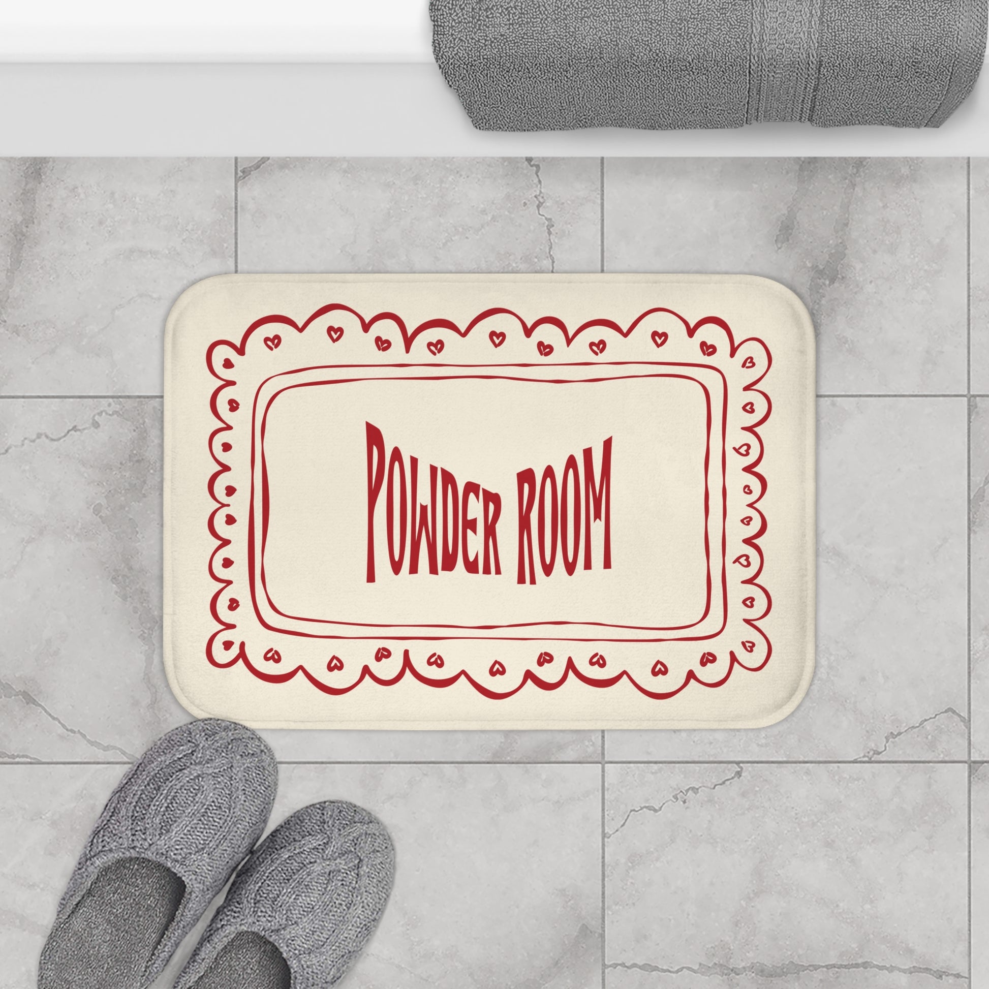 Beige bath mat with red 'Powder Room' text on a marble floor with slippers and a towel.