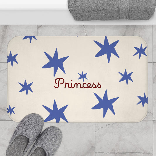 A rectangular bath mat with rounded corners in a bathroom setting, featuring a cream background with blue star patterns and the word 'Princess' in red script. Gray slippers and a folded gray towel are placed nearby on a light marble tile floor.