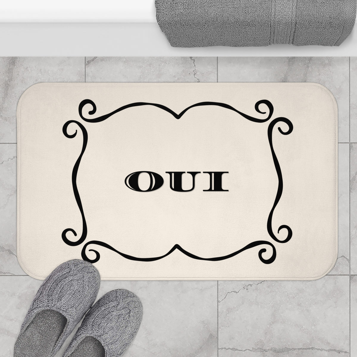 A rectangular bath mat with rounded corners in a warm cream color, featuring a black ornate scroll border and the word 'OUI' in the center. The mat is shown on a light gray marble tile floor, with gray knit slippers at the bottom left and a folded gray towel at the top.