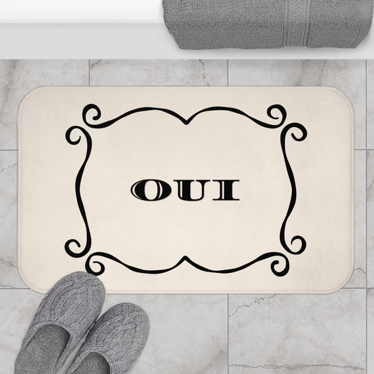 A rectangular bath mat with rounded corners in a warm cream color, featuring a black ornate scroll border and the word 'OUI' in the center. The mat is shown on a light gray marble tile floor, with gray knit slippers at the bottom left and a folded gray towel at the top.