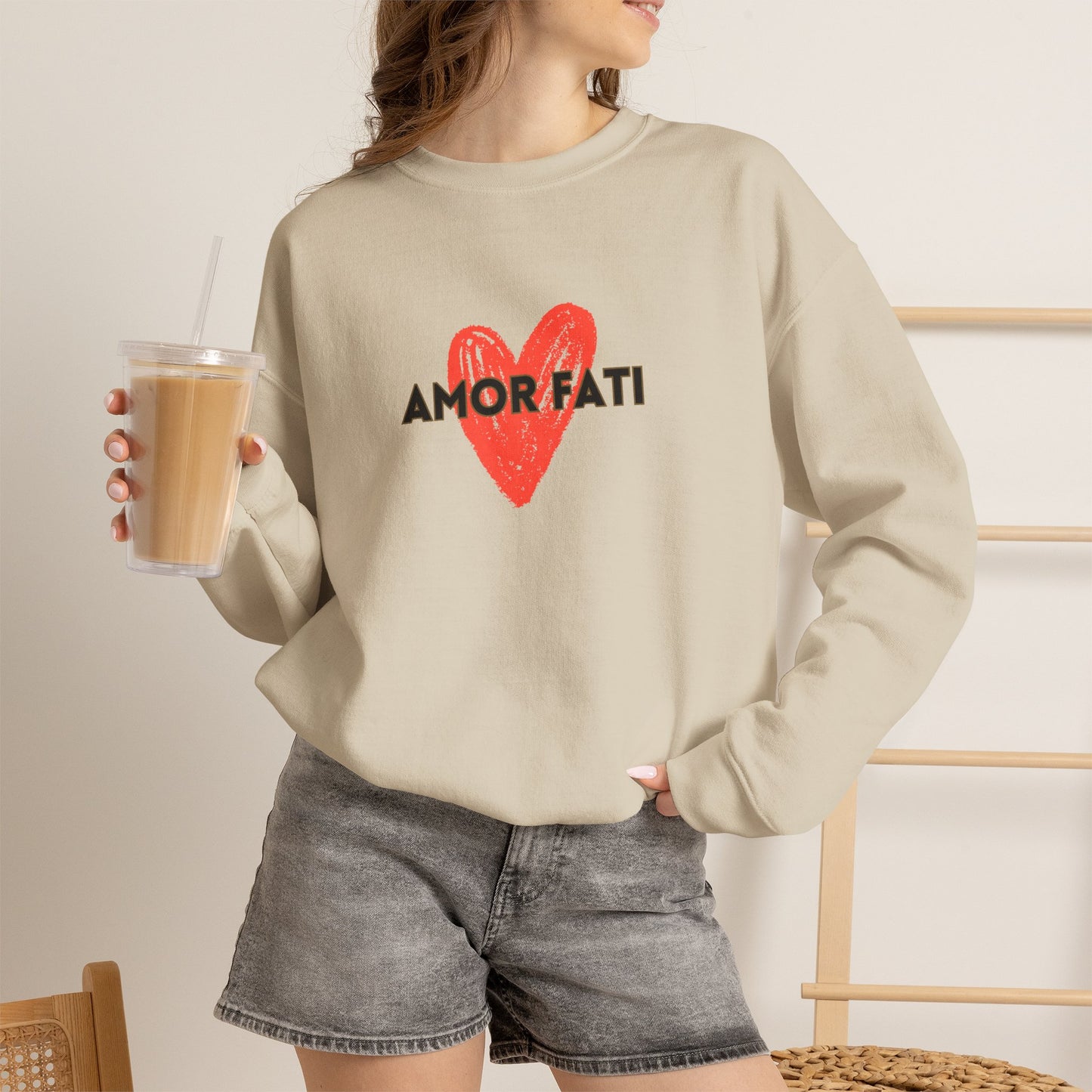 A woman wearing a beige crewneck sweatshirt with a red heart and black 'AMOR FATI' text graphic, paired with gray denim shorts and holding an iced coffee.