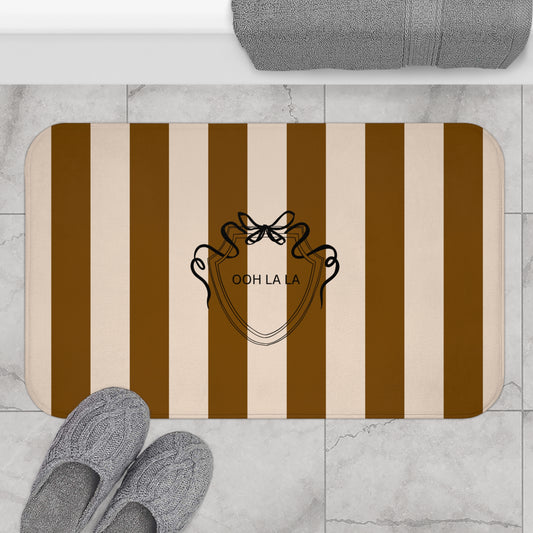 Striped bath mat with a logo and text on a marble floor with slippers and a towel.