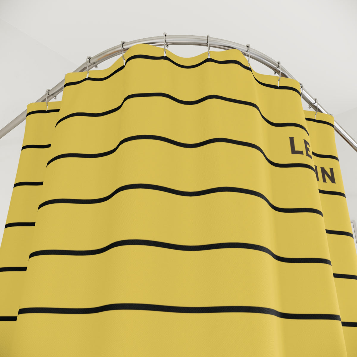 A yellow shower curtain with horizontal black wavy stripes and partial black text on the right side, hanging from a curved silver metal rod against a plain light background.