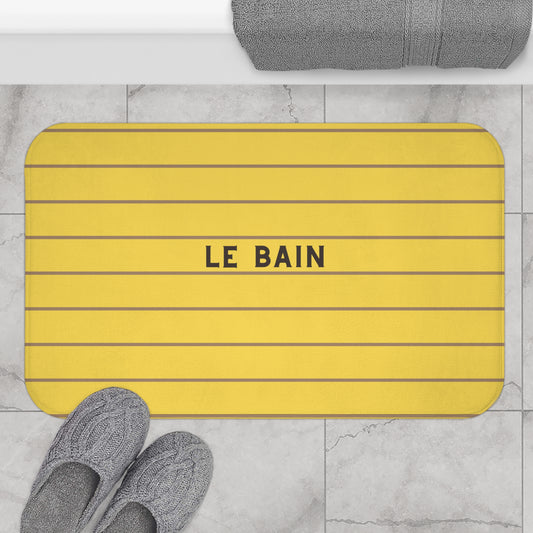 A yellow rectangular bath mat with thin dark horizontal stripes and the words 'LE BAIN' in the center, shown on a light marble floor with gray towels and slippers nearby.
