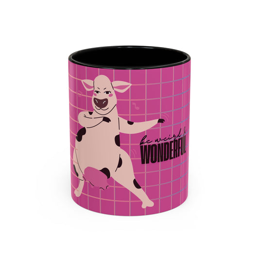 A ceramic mug with a pink and purple grid pattern featuring a cartoon pig character and the text 'be weird. WONDERFUL.