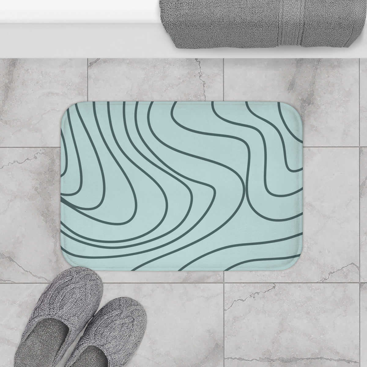 A rectangular bath mat with rounded corners featuring a light blue background and dark gray wavy contour-line pattern, shown on a gray marble-look tile floor in a bathroom setting with gray slippers and a folded towel visible.