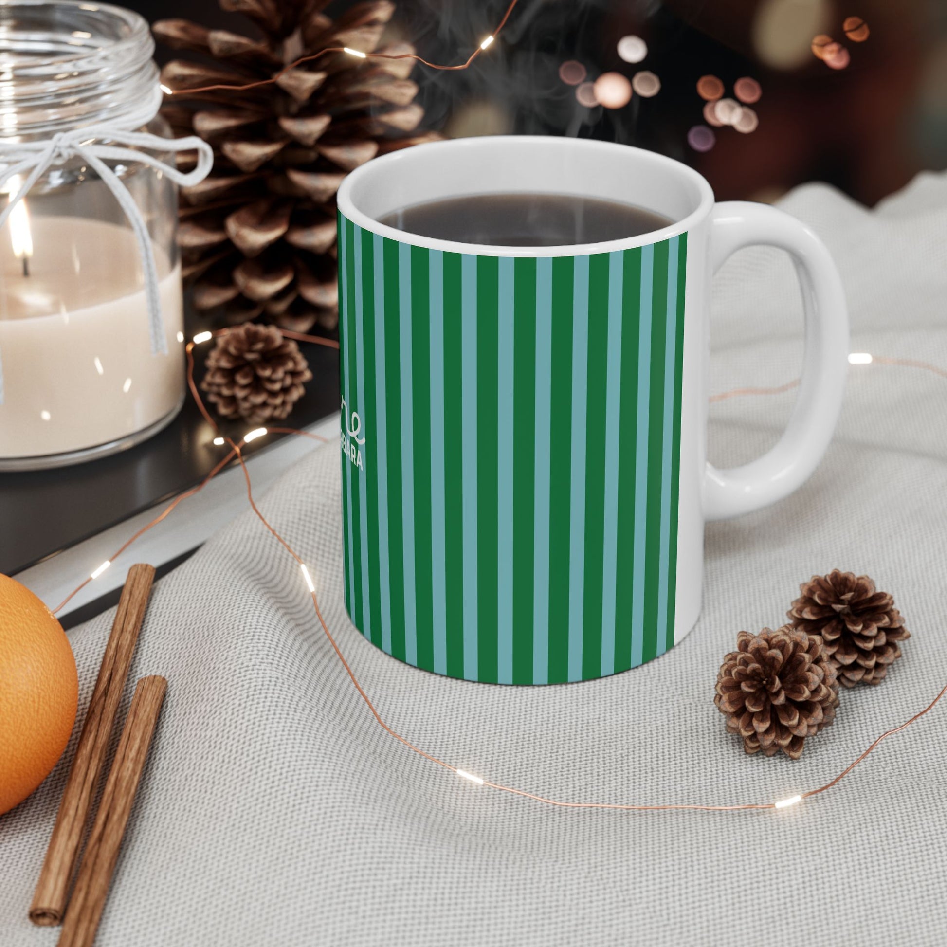 A white ceramic mug with green vertical stripes, filled with a hot beverage and set on a light fabric surface with pinecones, fairy lights, cinnamon sticks, an orange, and a candle in the background.