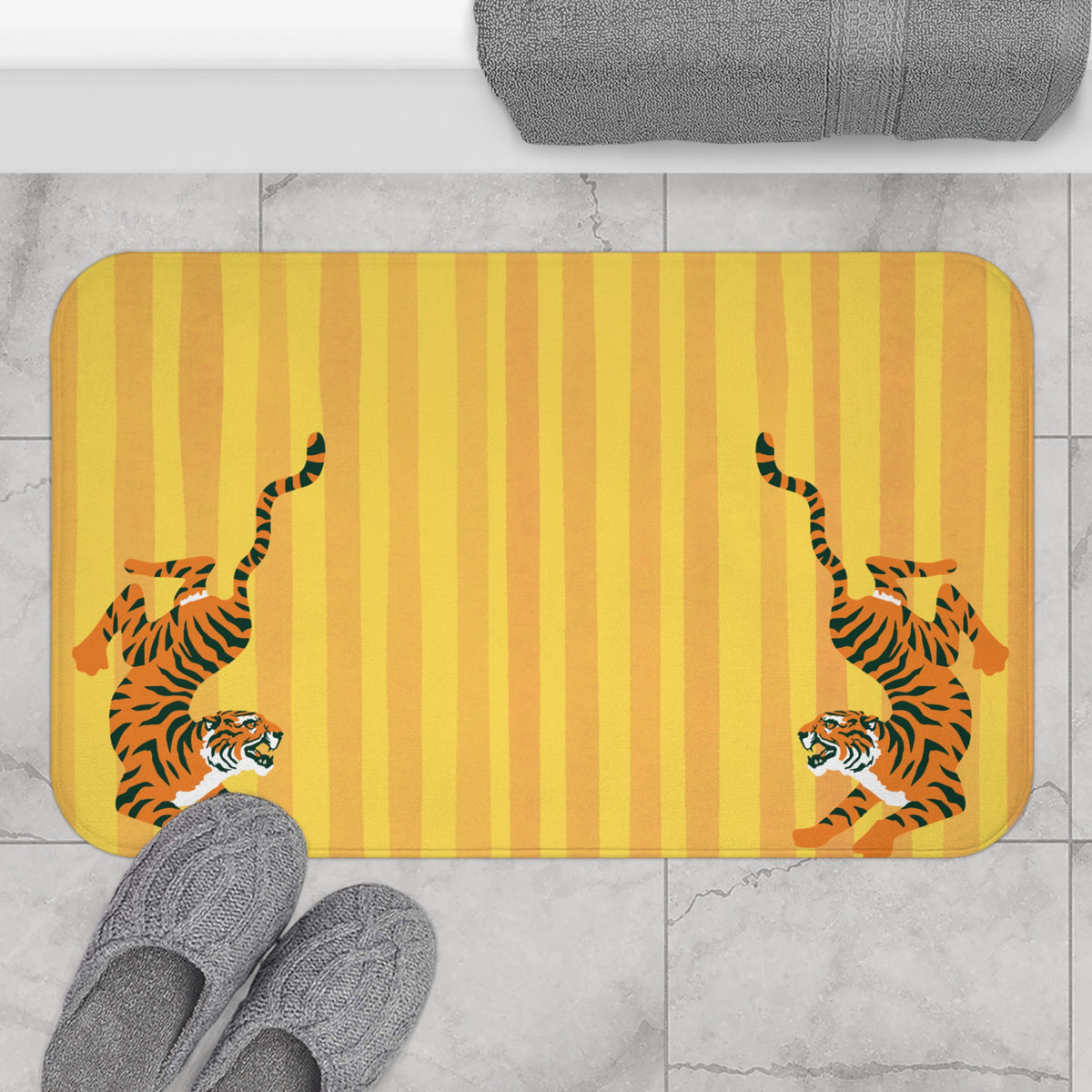 A rectangular bath mat with rounded corners featuring two cartoon tigers on a yellow and orange striped background, shown on a light marble floor with gray towels and slippers nearby.