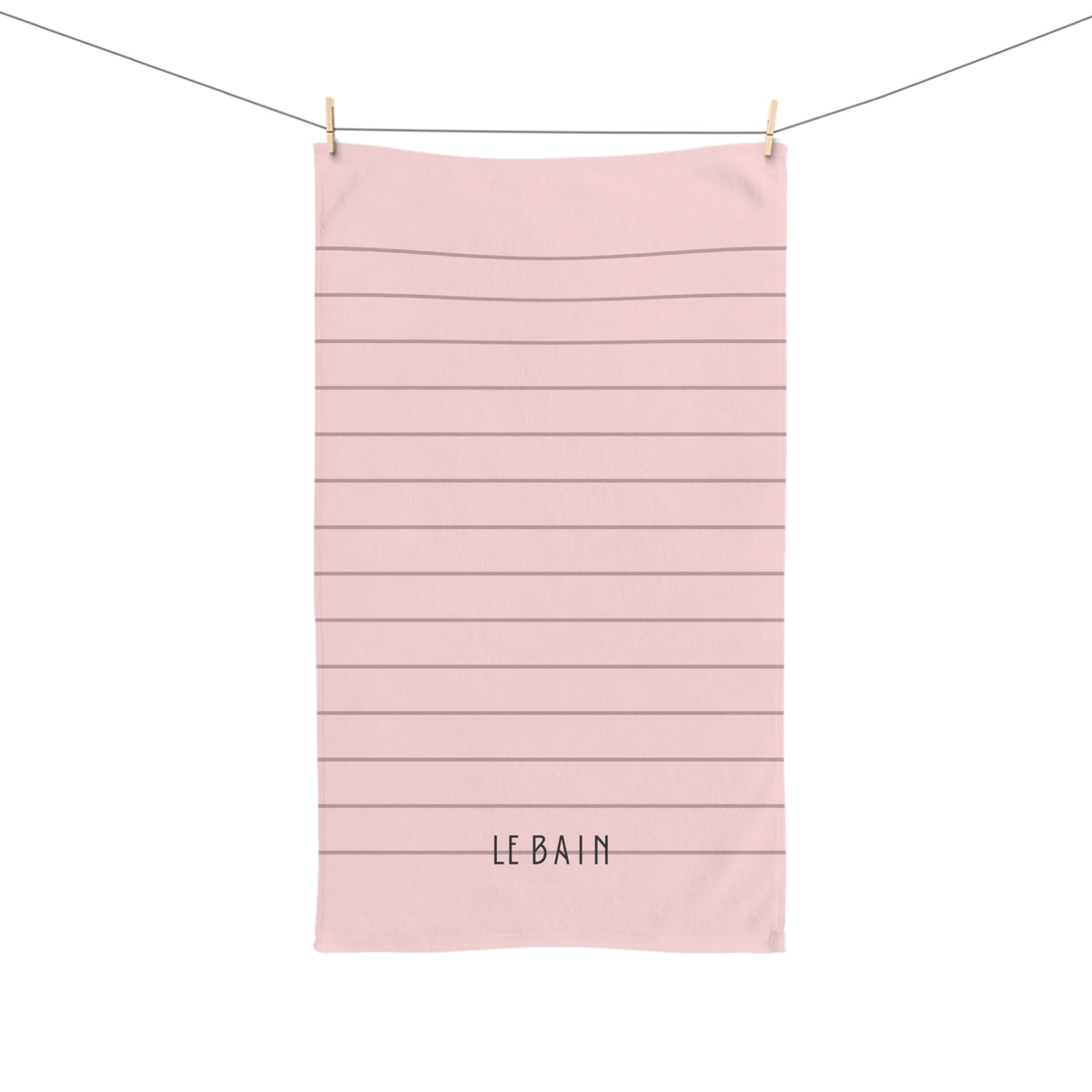 A light pink kitchen towel with horizontal dark gray stripes and the text 'LE BAIN' printed at the bottom, hanging from a clothesline with two wooden clips against a white background.
