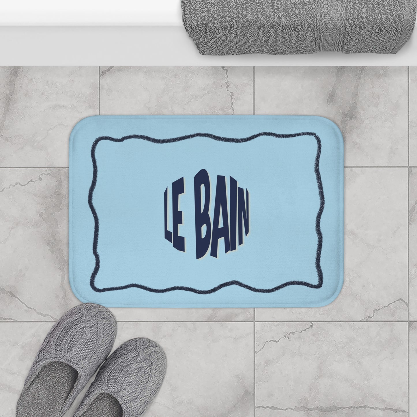 Light blue bath mat with 'LE BAIN' text on a tiled bathroom floor.